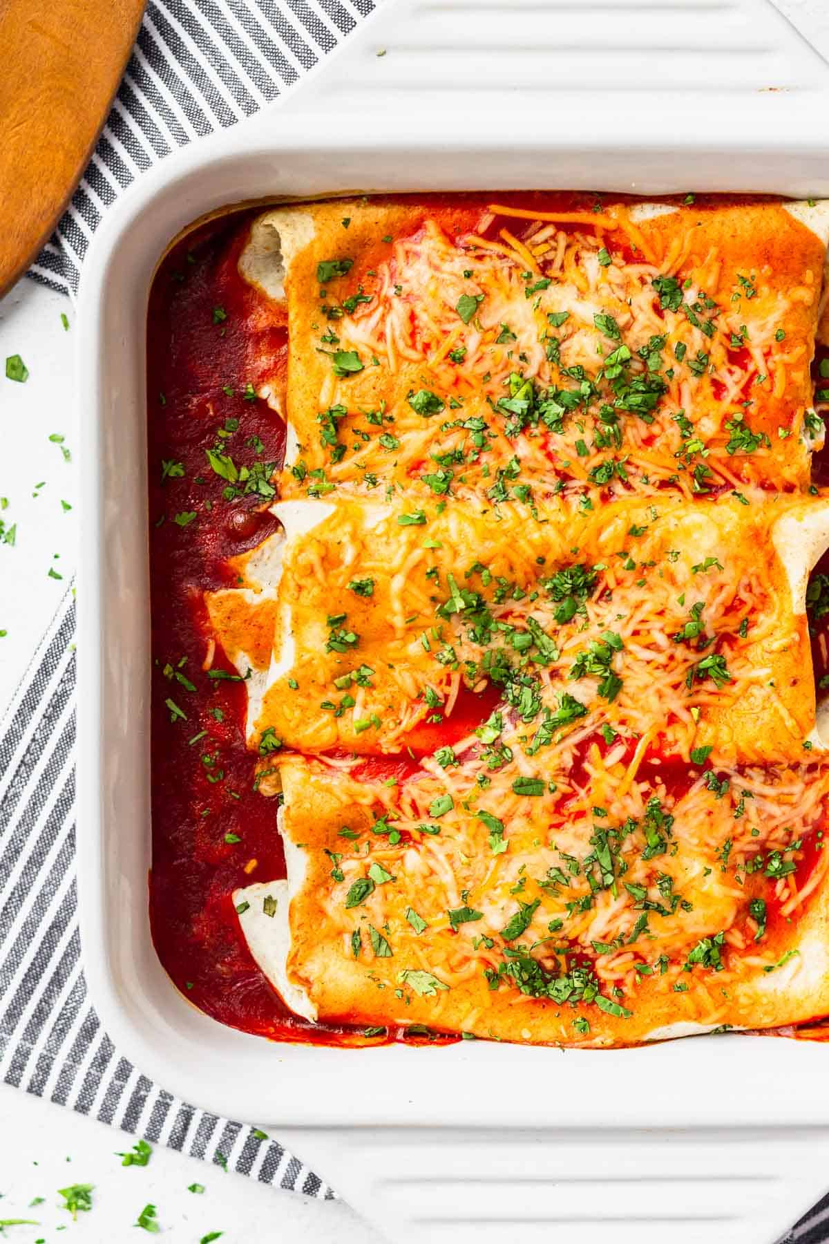 Close up of 3 large chicken enchiladas in a white casserole dish, with part of a striped towel under it