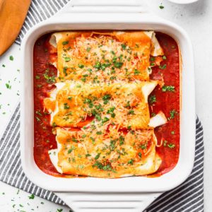 Baked cheesy chicken enchiladas in a white, square baking dish with a striped towel and server next to it