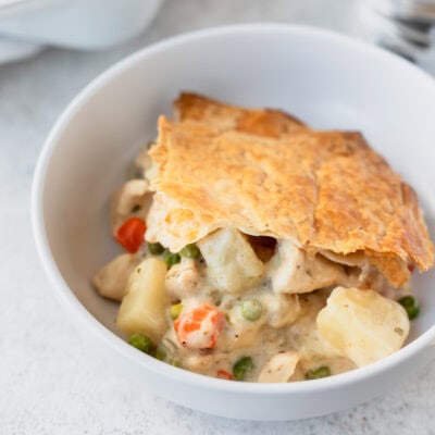 A serving of creamy chicken pot pie topped with puff pastry and filled with chicken, potatoes, carrots and peas in a white bowl.