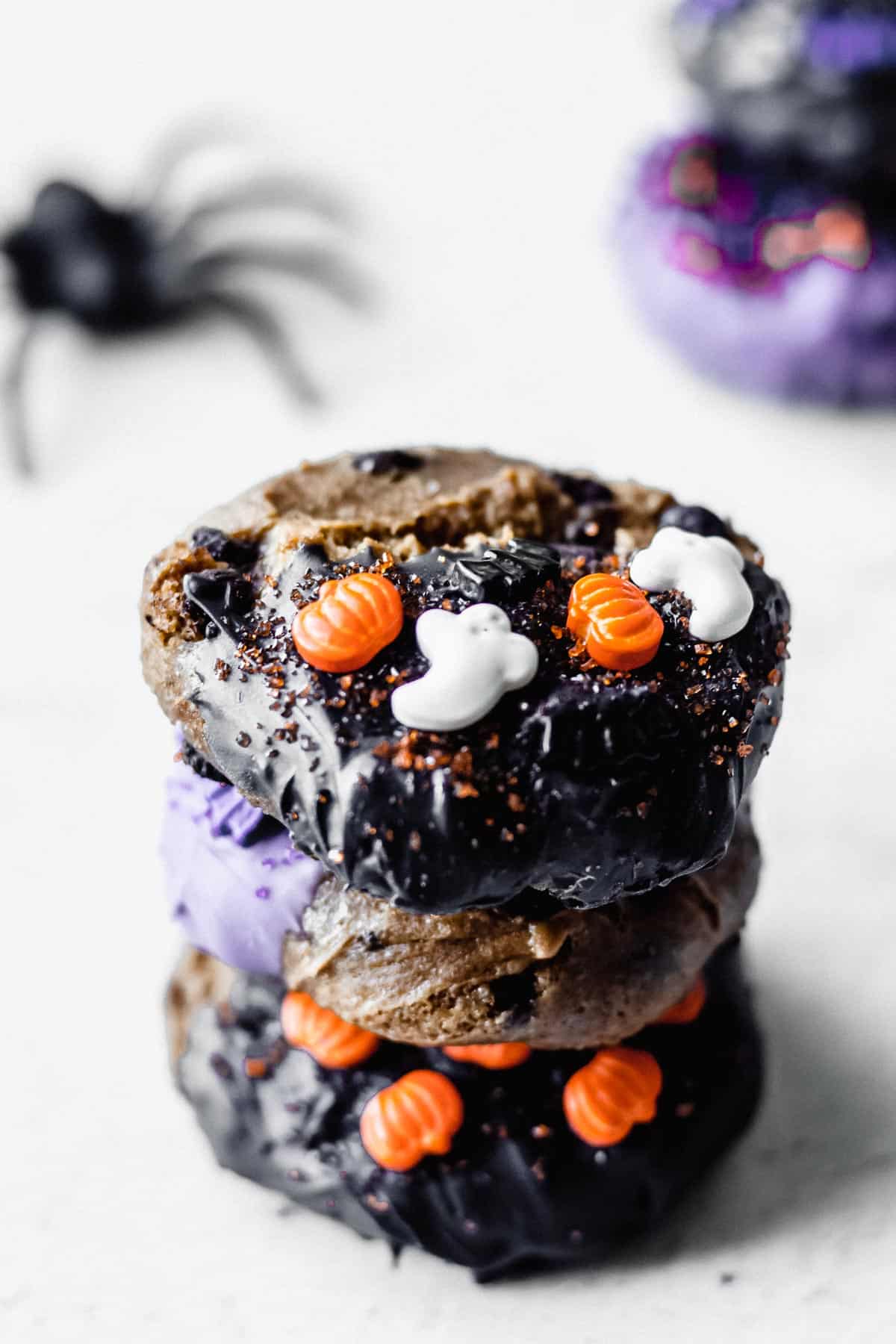 3 chocolate chip cookies with halloween sprinkles on them in a stack with a toy spider and more cookies in the background