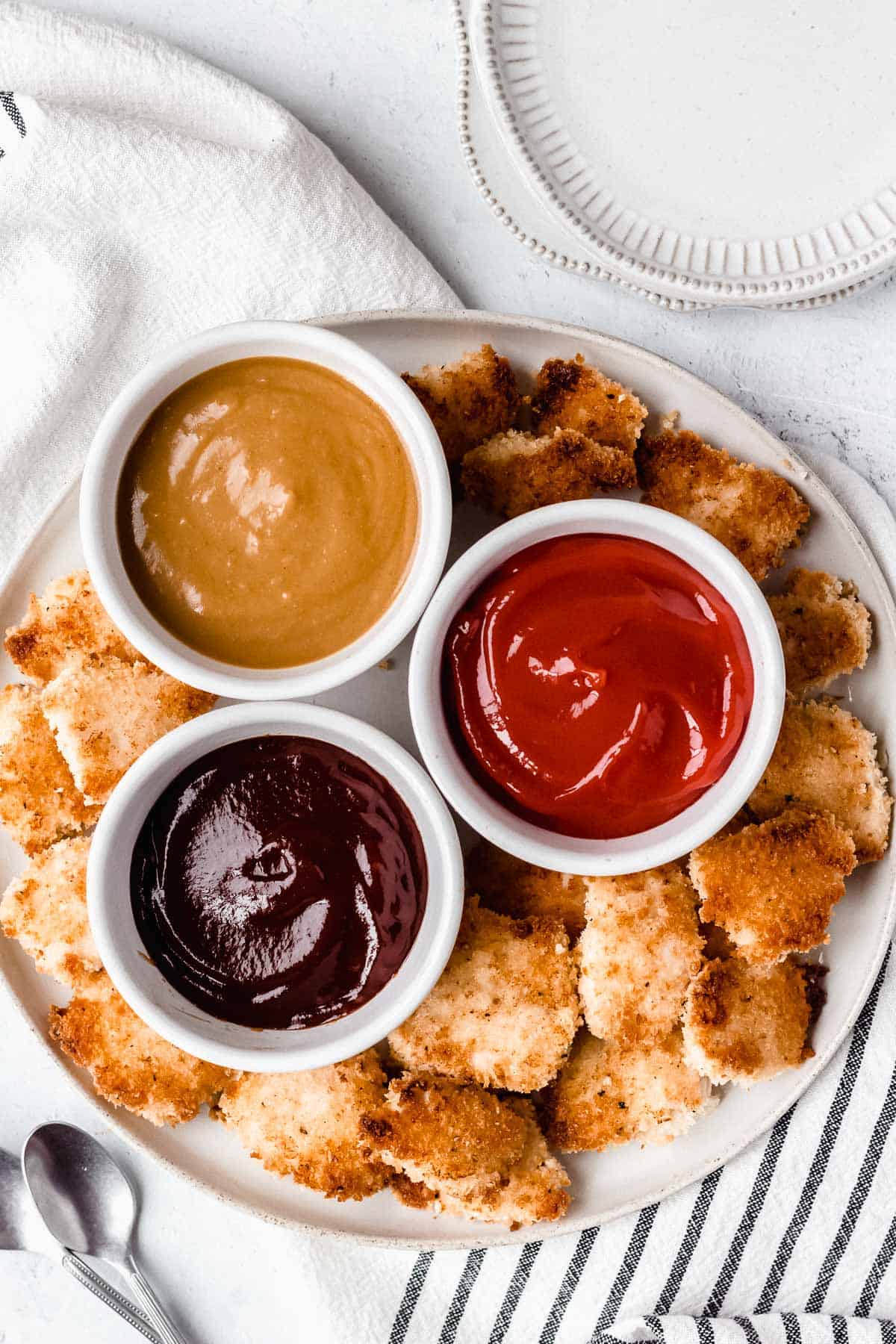 Overhead of mayonnaise chicken nuggets on a white plate with 3 white bowls of different sauces and a striped towel and small white plates around it on a white background