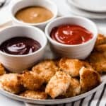 Homemade chicken nuggets on a plate with 3 whites bowls of dipping sauces