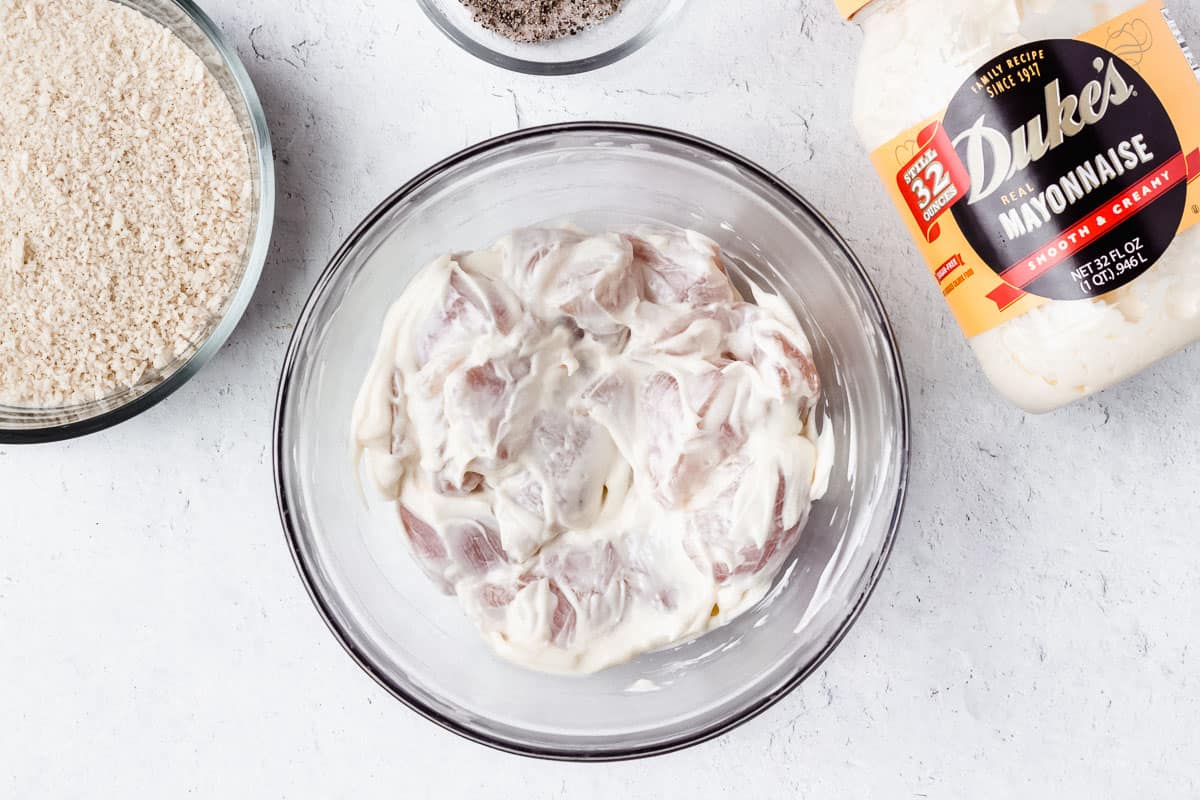 Chunks of chicken coated in mayonnaise in a glass bowl with a bowl of bread crumbs and container of mayonnaise around it