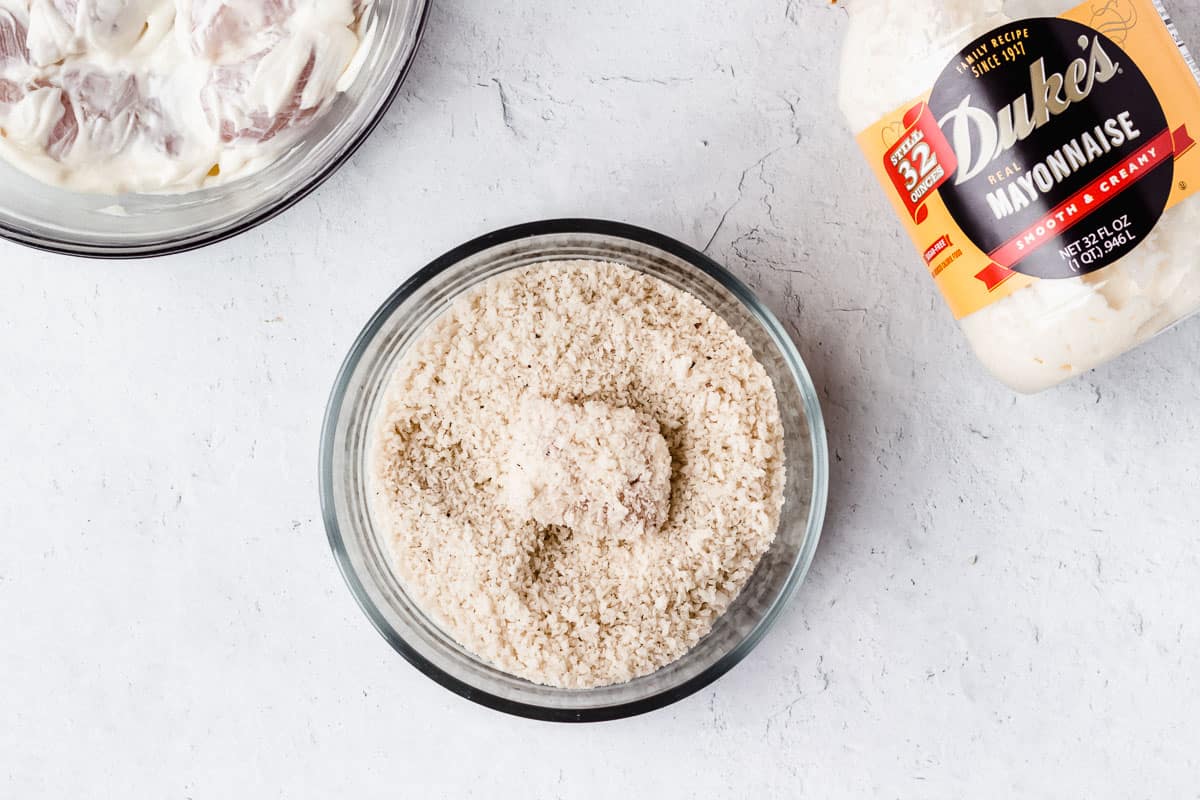 A chunk of chicken coated with panko bread crumbs in the bowl of crumbs with a bowl of chicken and container of mayonnaise in the background