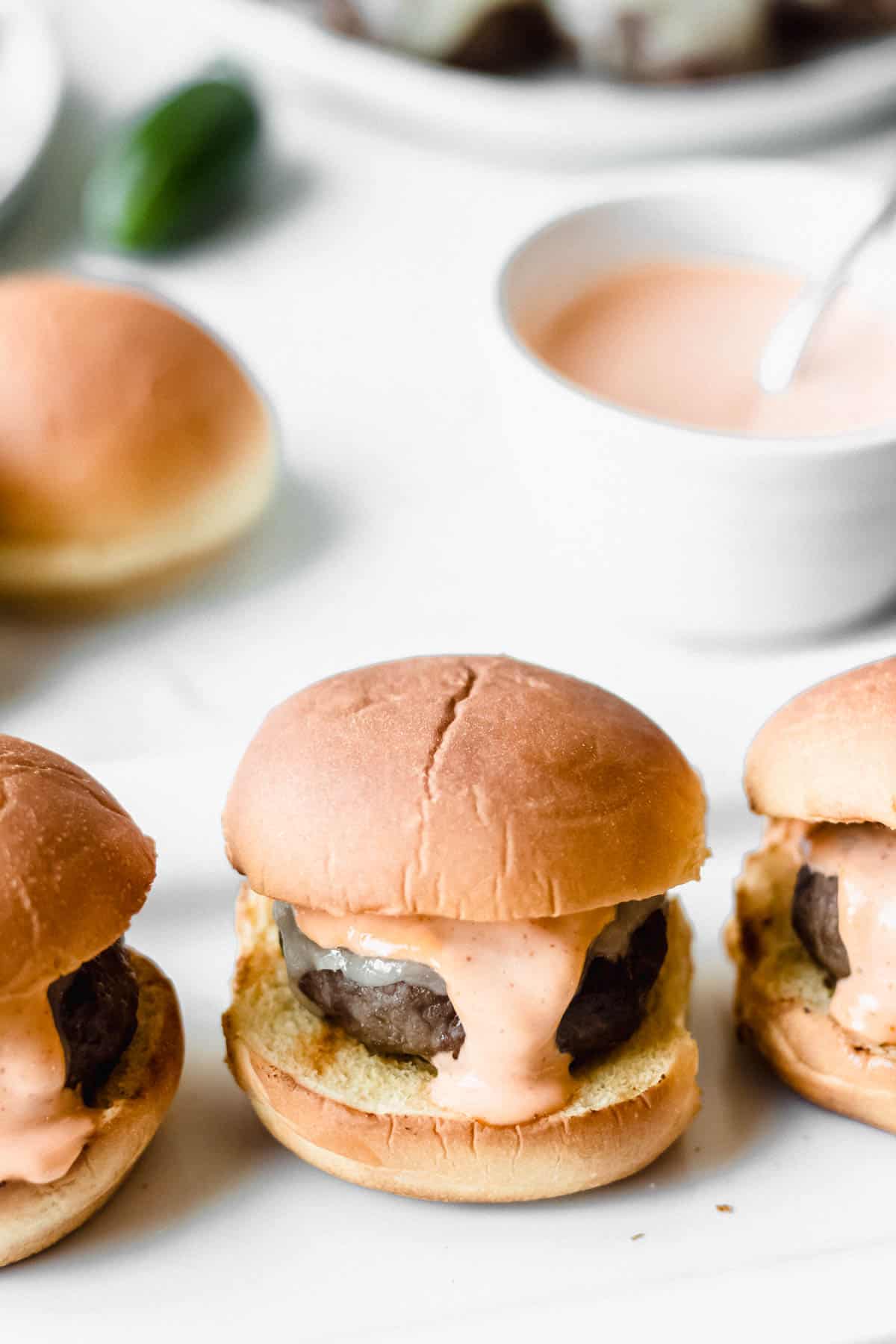 Close up of spicy beef sliders with a bowl of spicy mayo, buns, and a plate of burgers partially showing in the background
