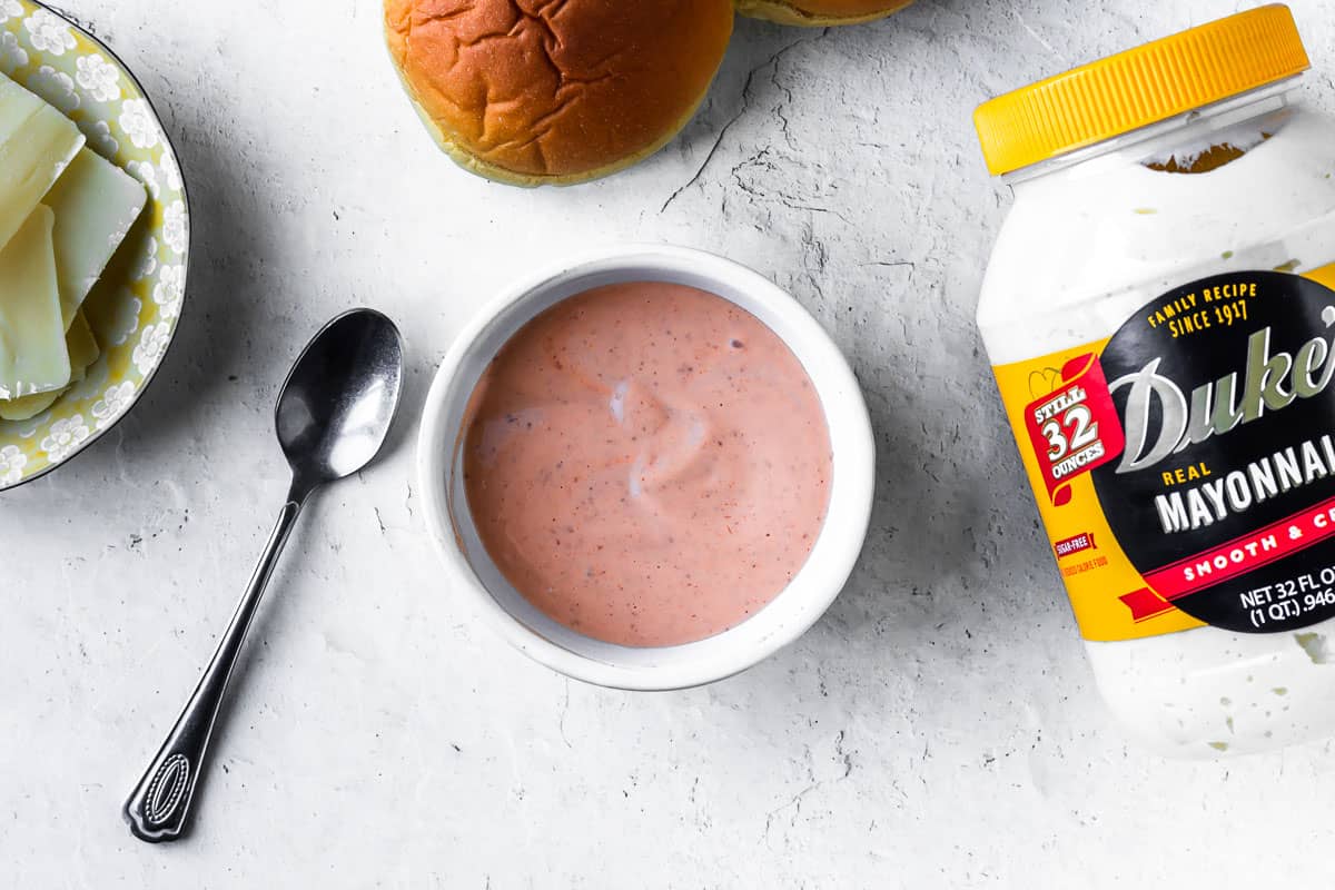 Spicy mayonnaise in a white bowl with a spoon next to it and other ingredients in the background on a white backdrop