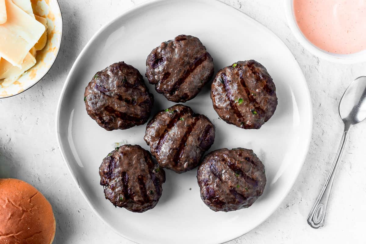 8 grilled beef sliders on a plate with other ingredients around it on a white background