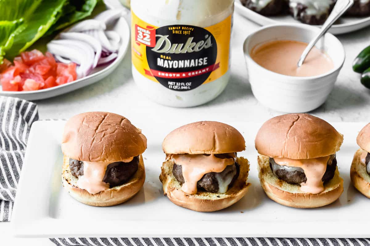 4 spicy beef sliders on a white plate with duke's mayonnaise, burger toppings, and spicy mayo in the background