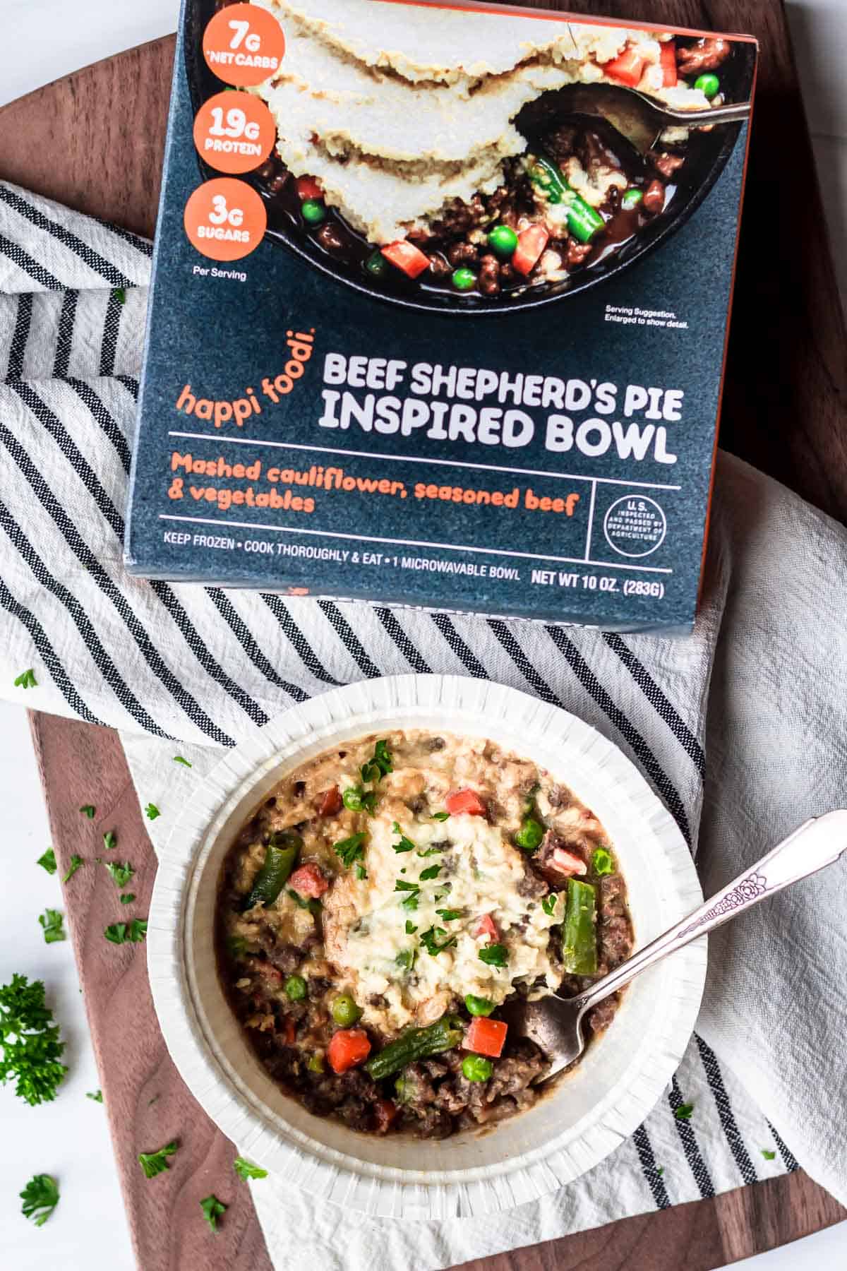 Happi Foodi Beef Shepard's Pie Inspired Bowl box and meal on a wood background with a striped napkin, fork, and parsley