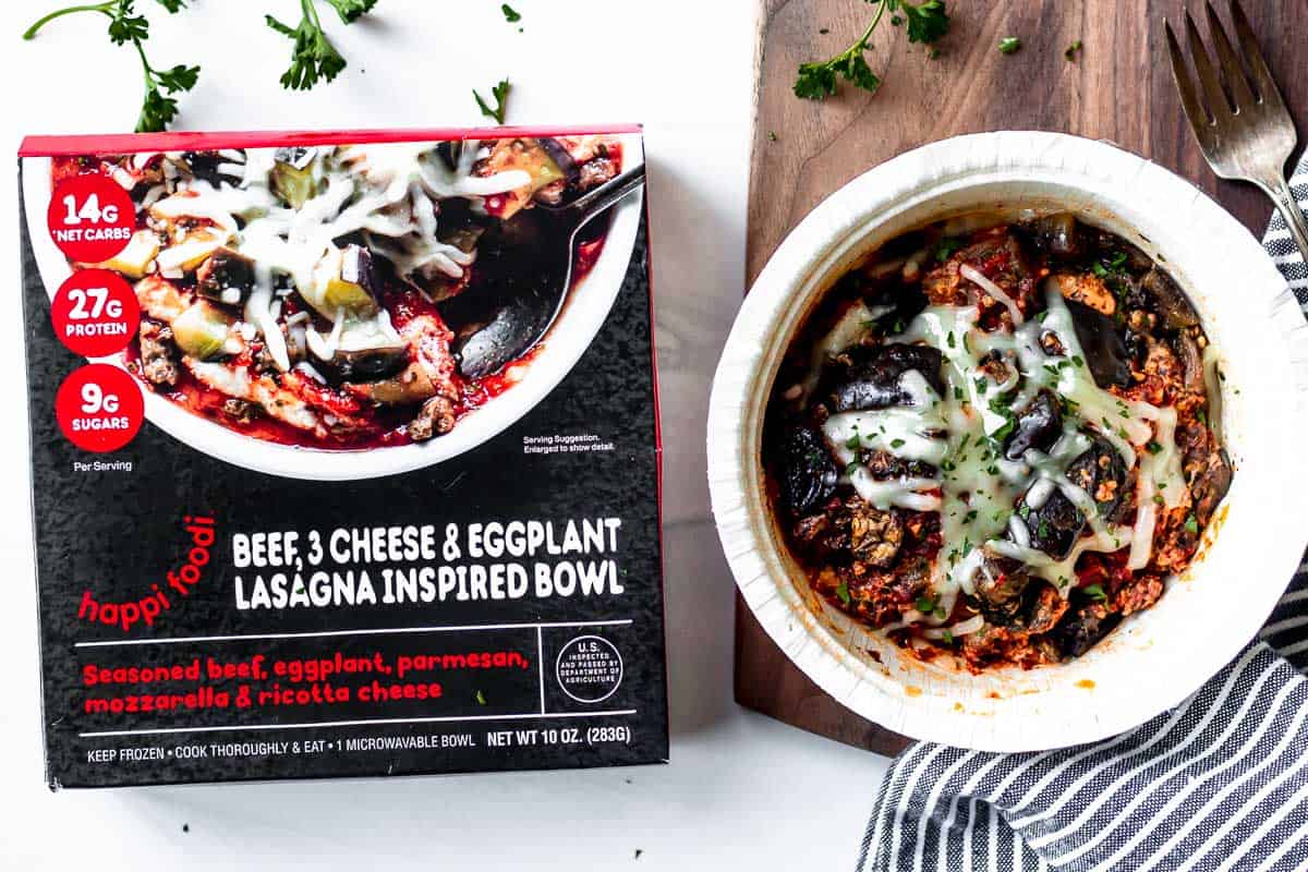 Happi Foodi's Beef, 3 Cheese, & Eggplant Lasagna Inspired Bowl next to the package on a wood board and white background with a striped napkin