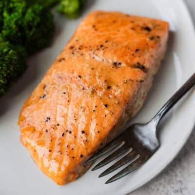 A maple bourbon salmon fillet on a white plate with a fork and broccoli.