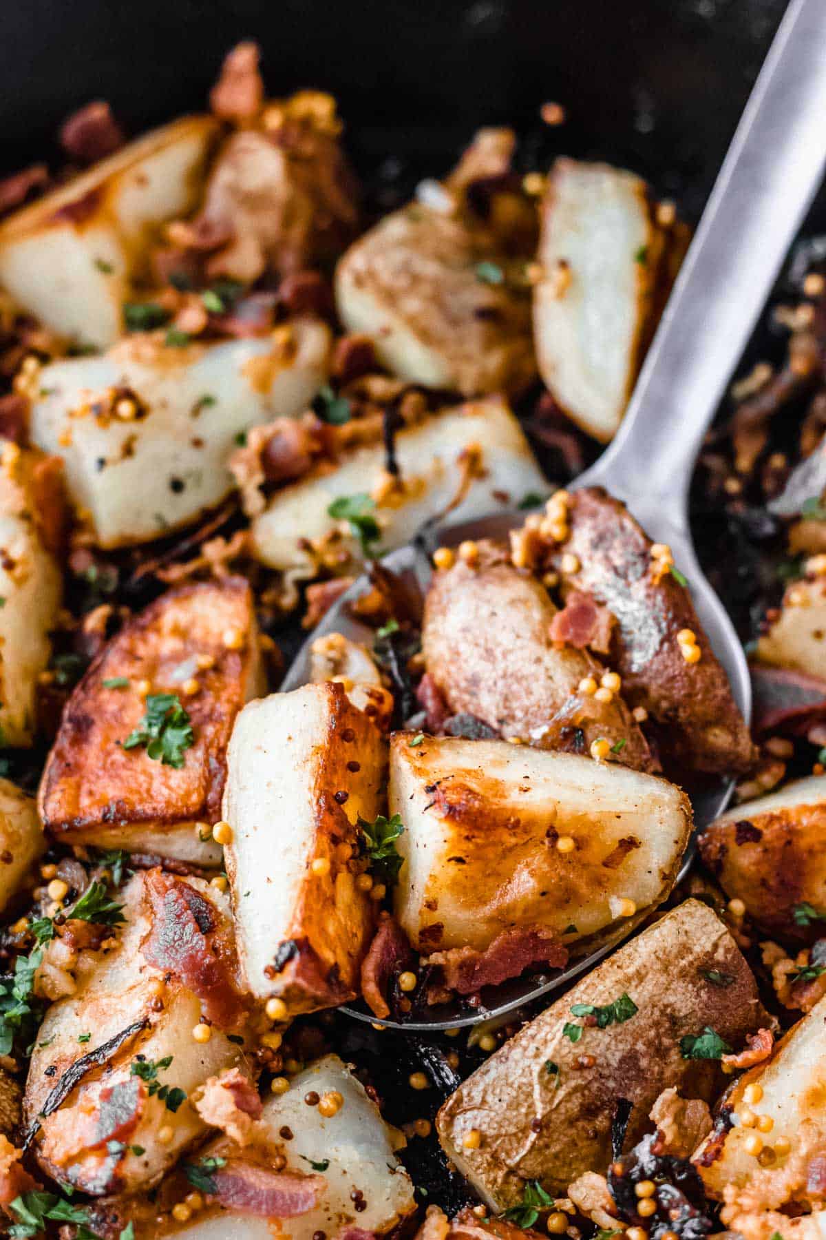 CLose up of a silver spoon with bacon roasted potatoes, caramelized shallots, mustard and parsley