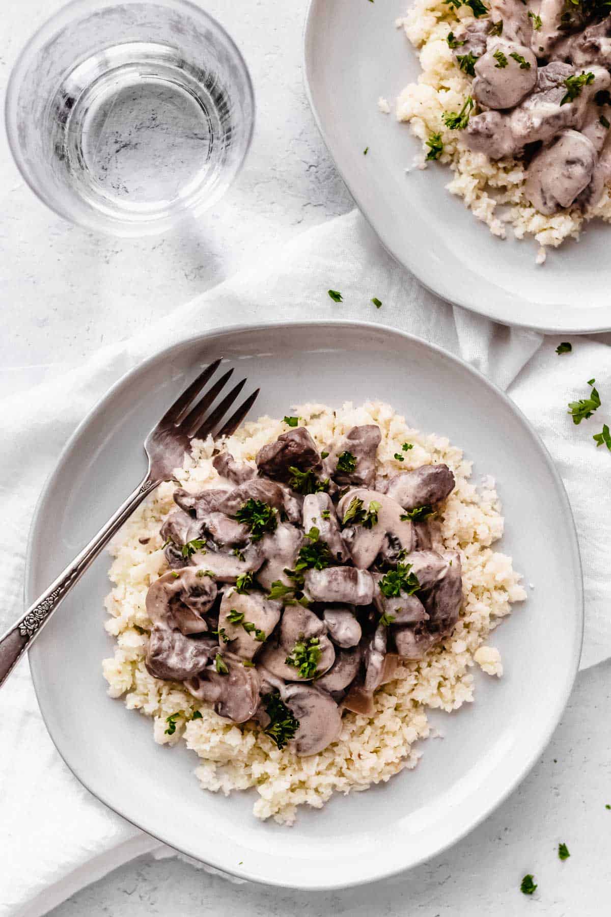 Overhead of keto beef stroganoff on a white plate with a second plate and a glass of water partially showing