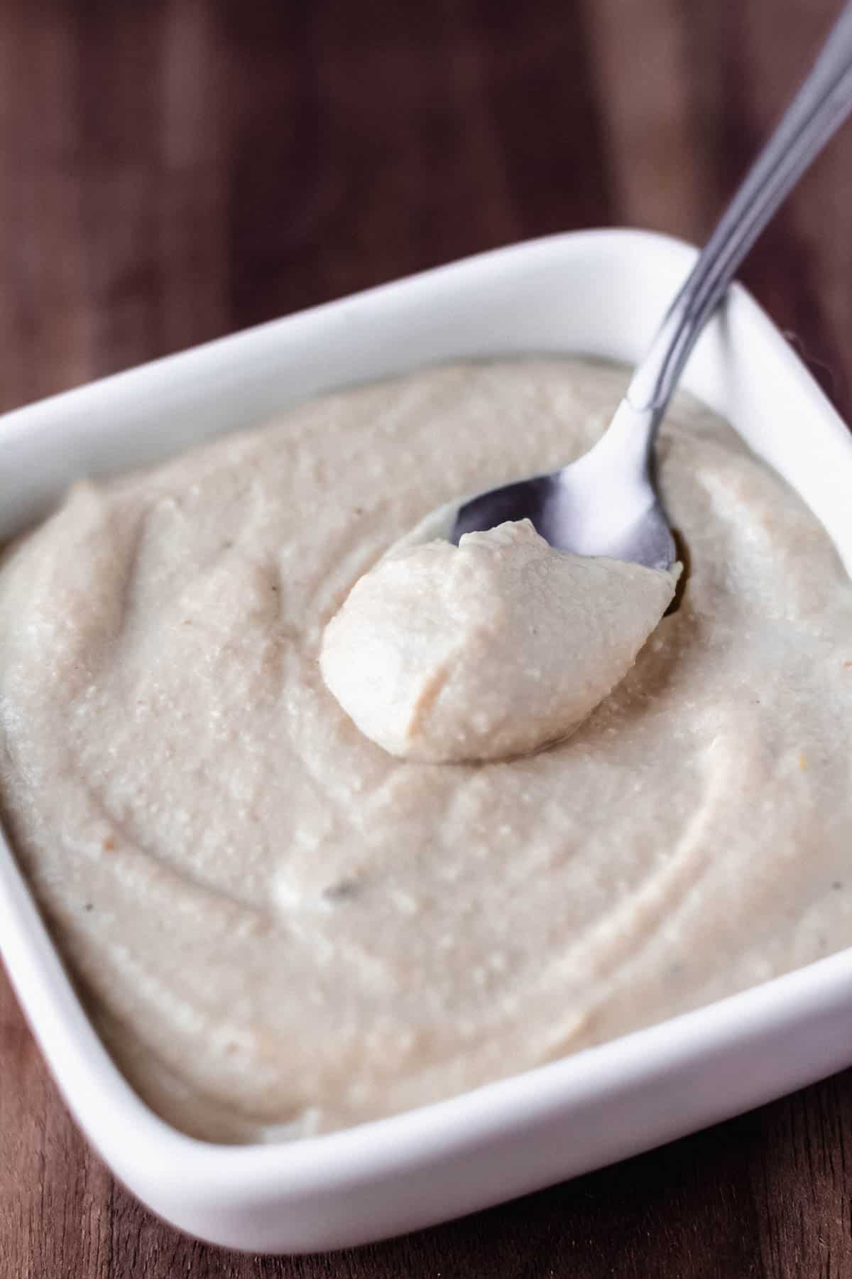 A spoon of cashew dip in a square dish of more dip over a white background