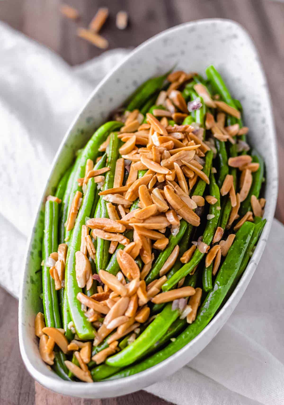 An oblong bowl filled with green beans and almonds over a white towel and wood board