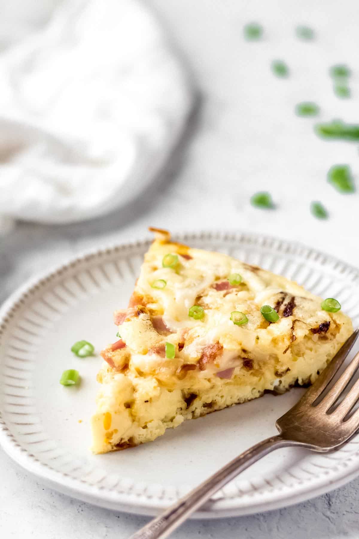 A slice of ham and cheese frittata on a small white plate with a fork and a white napkin and extra green onions in the background