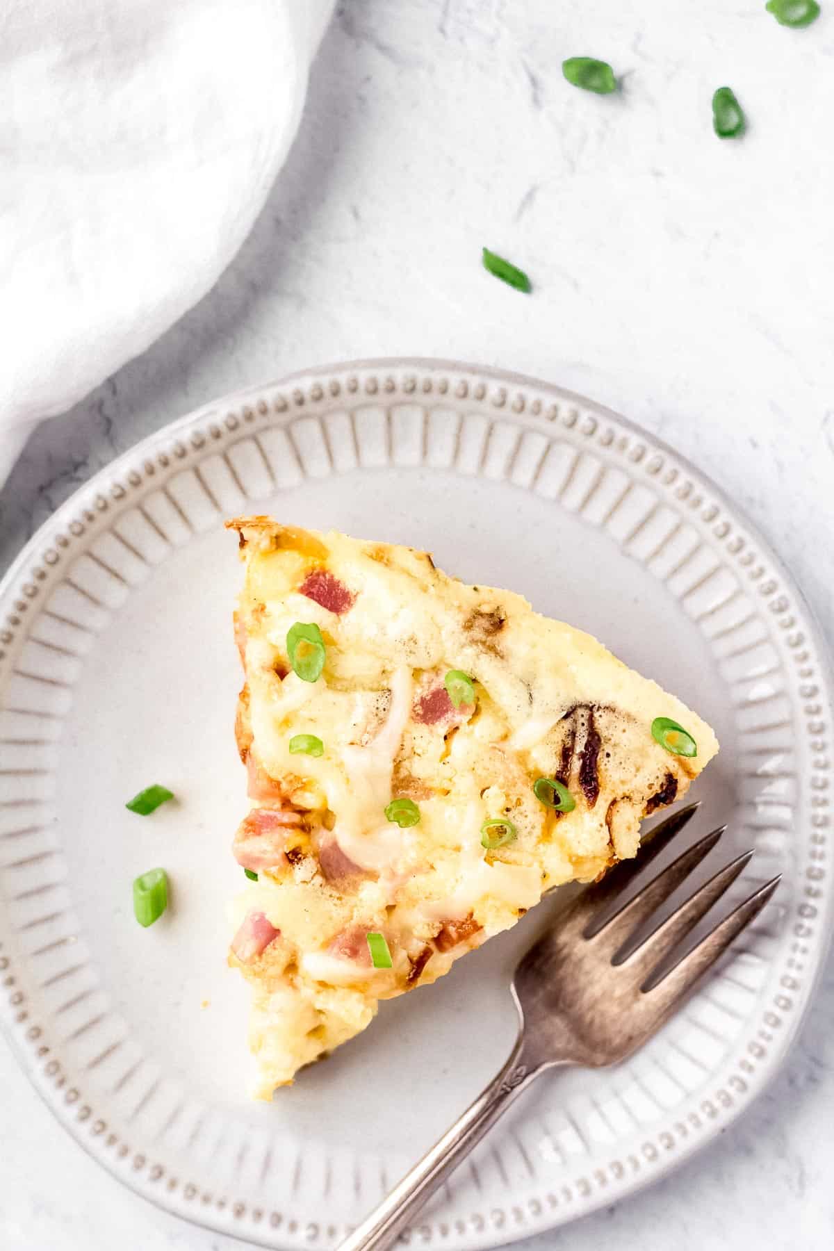 Overhead of ham and cheese frittata on a small white plate with a fork and a white napkin and slices of green onion in the background
