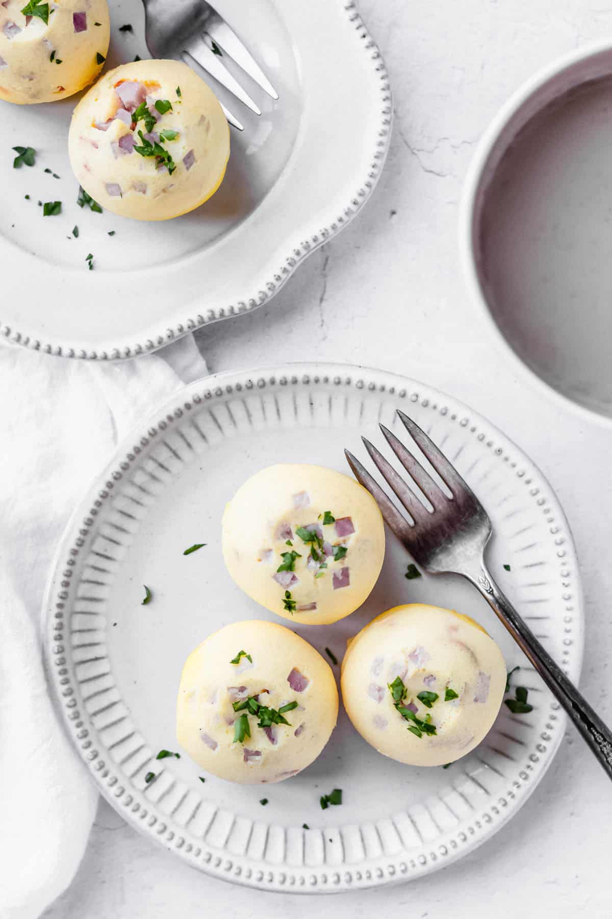 Overhead of 3 instant pot egg bites with ham on a white plate with a second plate of egg bites showing and part of a cup of coffee. There is also a fork on each plate and a white towel off to the side