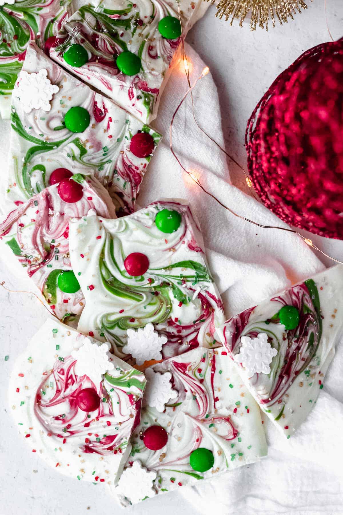 Pieces of Christmas bark laid on top of each other on a white napkin with twinkle lights and part of a red glittery tree around it