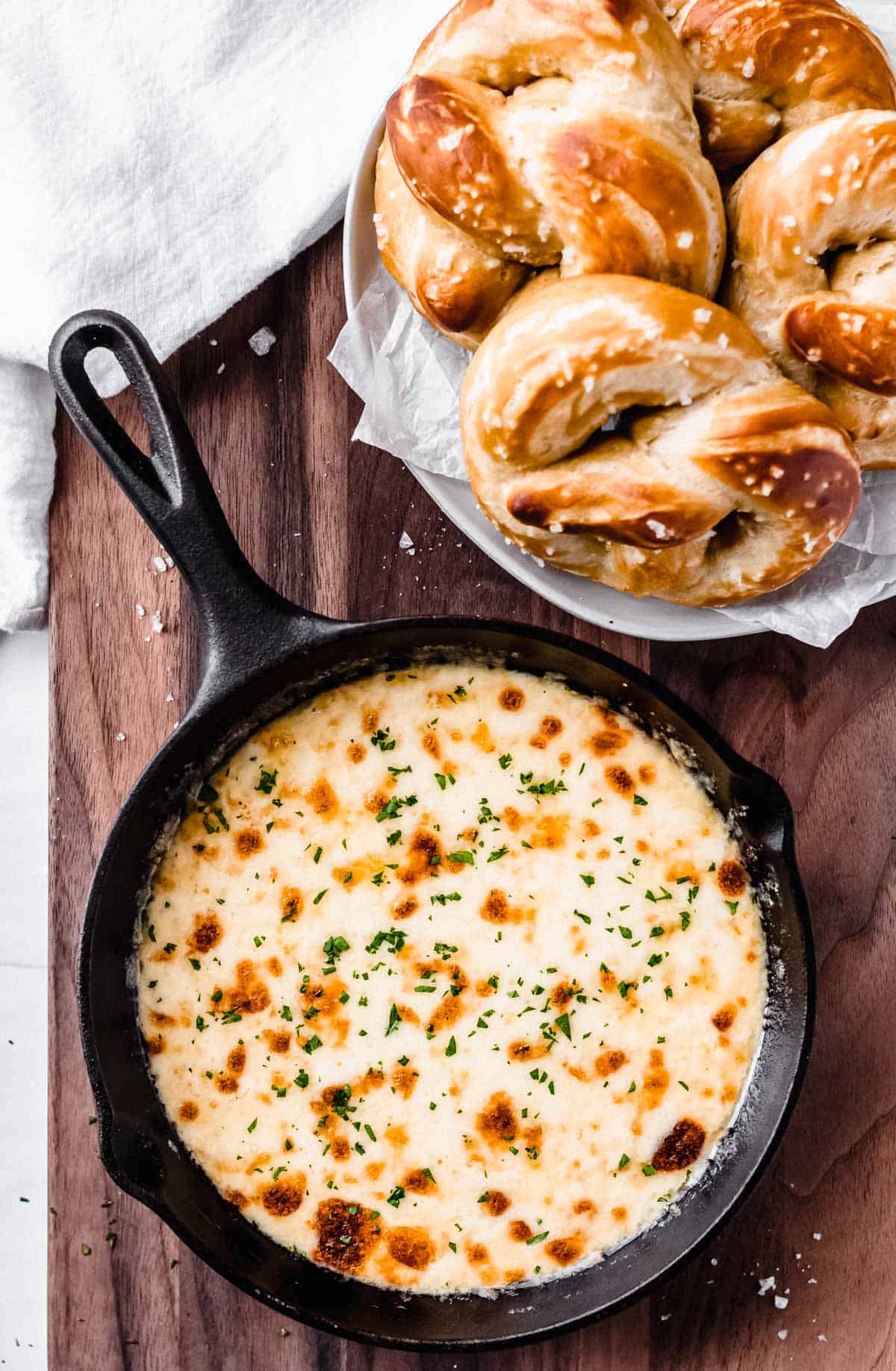 Overhead view of creamy white cheddar beer cheese dip in a black cast iron pan on a wood background with a plate of soft pretzels and a white towel behind it