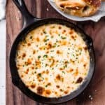 Overhead view of creamy white cheddar beer cheese dip in a black cast iron pan on a wood background with part of a plate of soft pretzels behind it