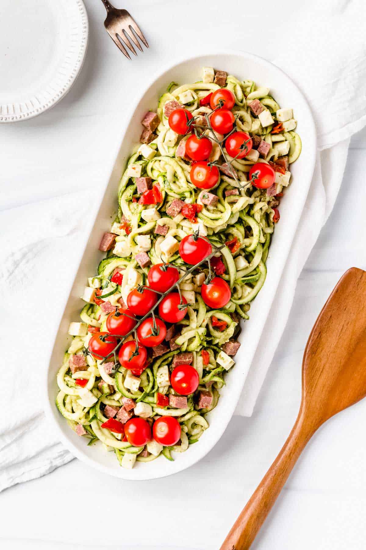 Overhead of keto pesto pasta salad in an oval serving dish on a white backdrop with a white towel, small white plate, fork and server around it