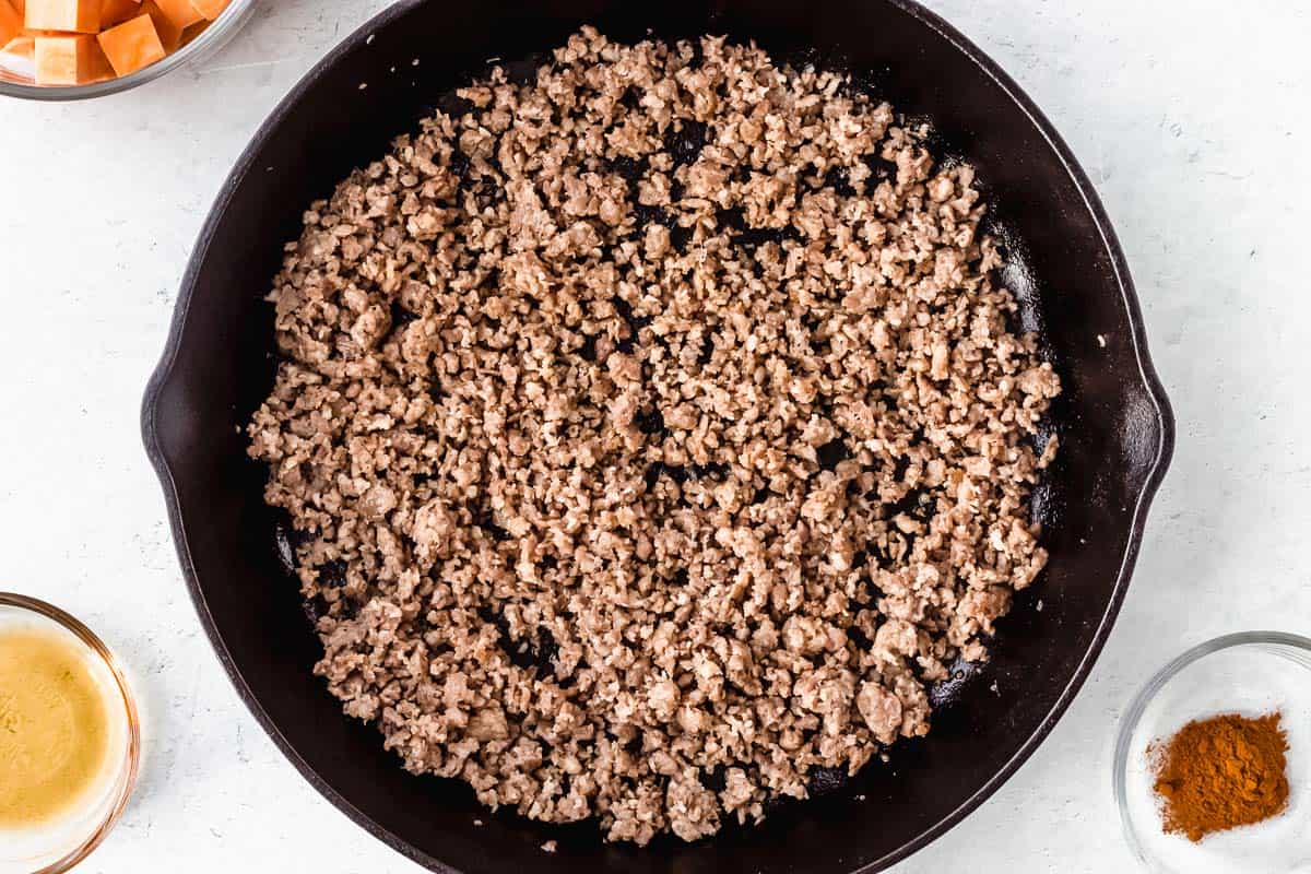 Crumbled sausage in a black cast iron skillet with other ingredients in glass bowls around it