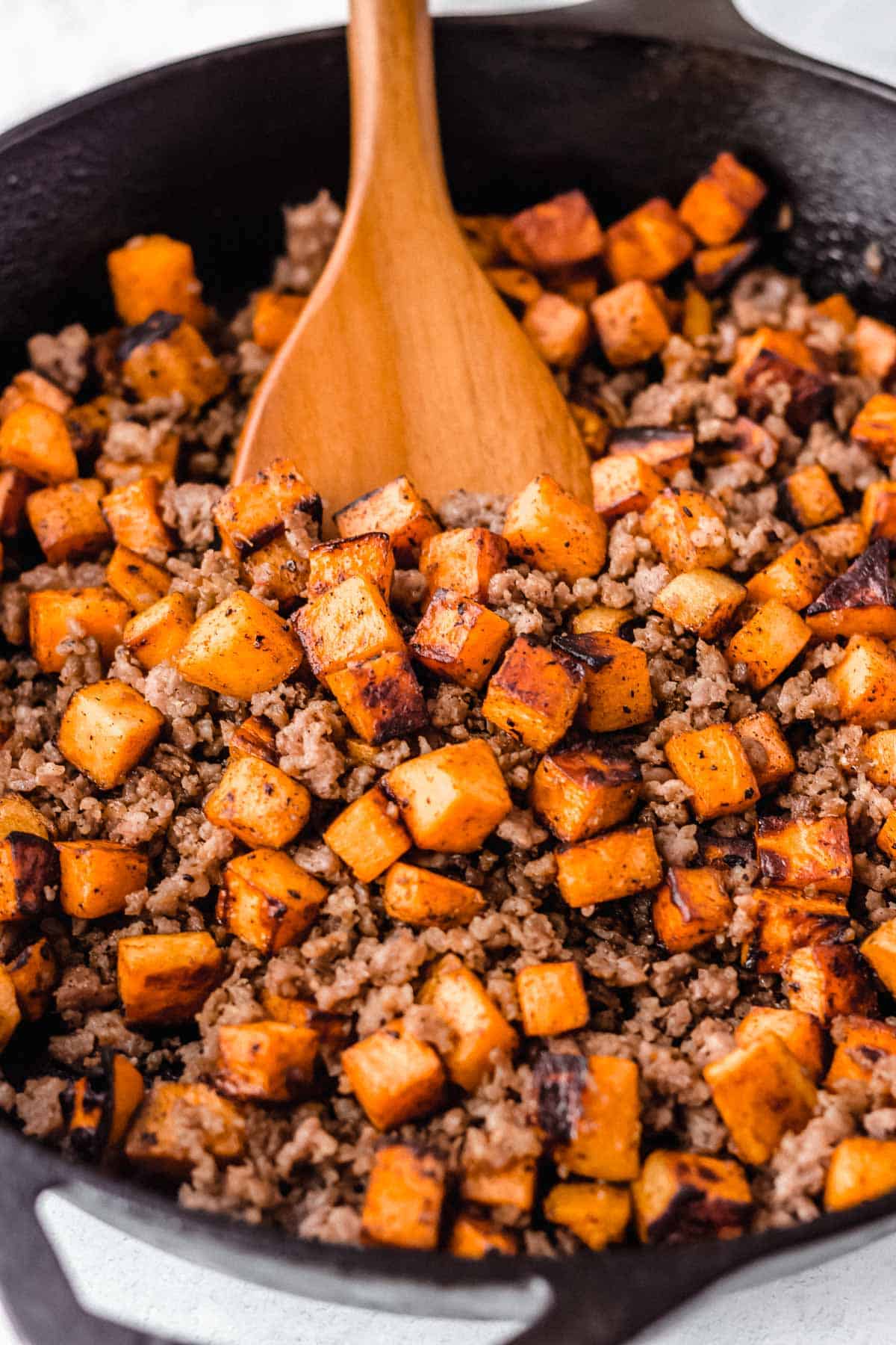 Cast iron pan full of maple sausage sweet potato hash with a wood server pushing up a scoop