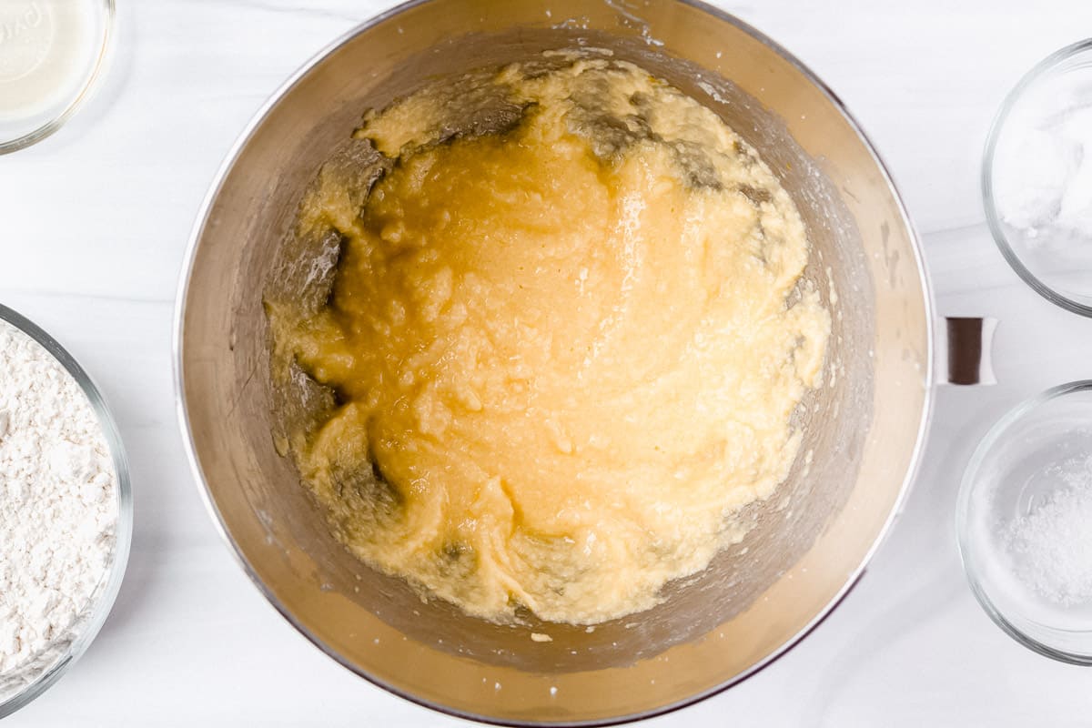 Cake batter with butter, sugar, eggs and vanilla blended together in a silver mixing bowl over a white background with bowls of additional ingredients around it
