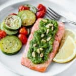 Baked Pesto Salmon on a white plate with lemon slices, zucchini and tomatoes