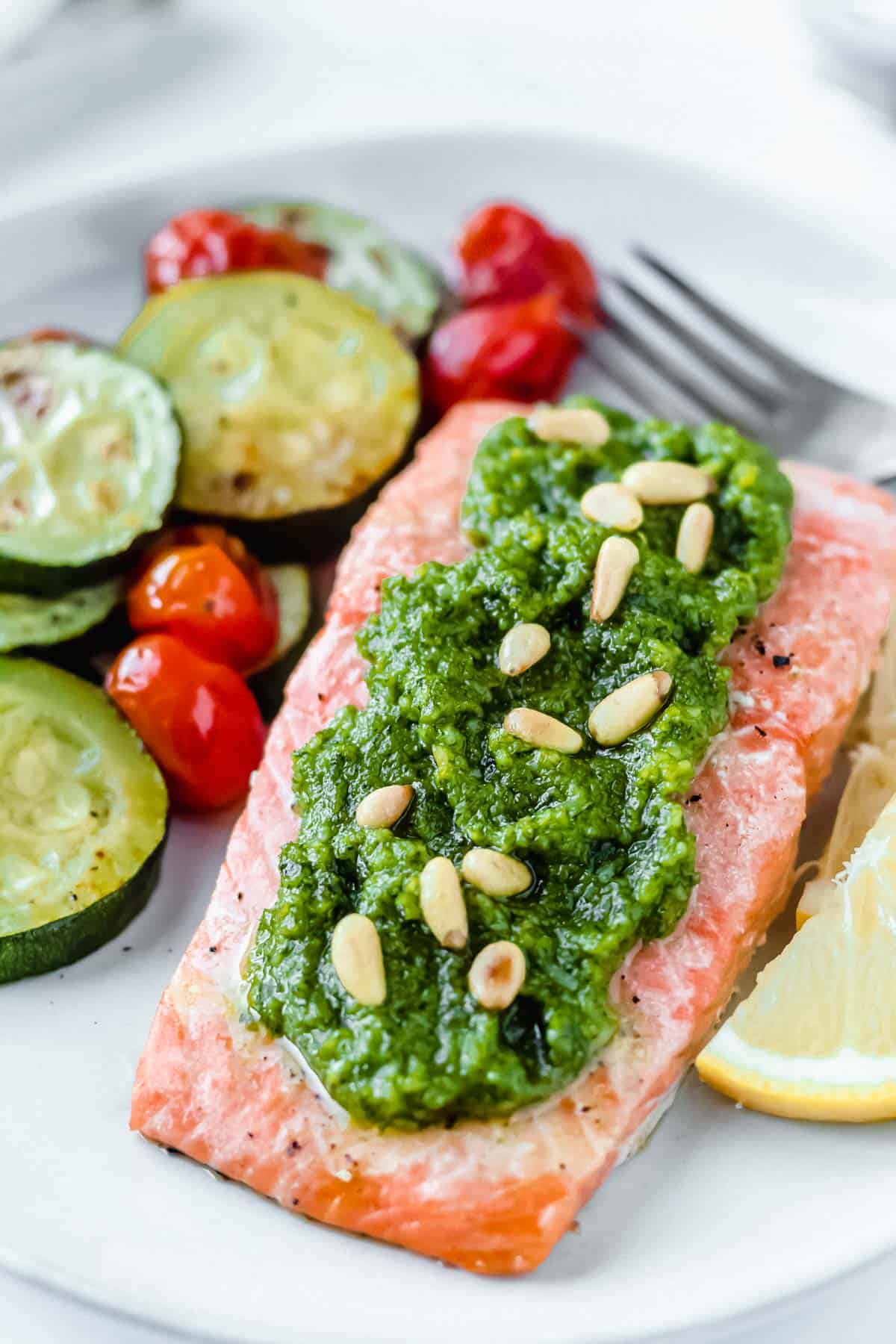 Baked Pesto Salmon on a white plate with lemon slices, zucchini and tomatoes