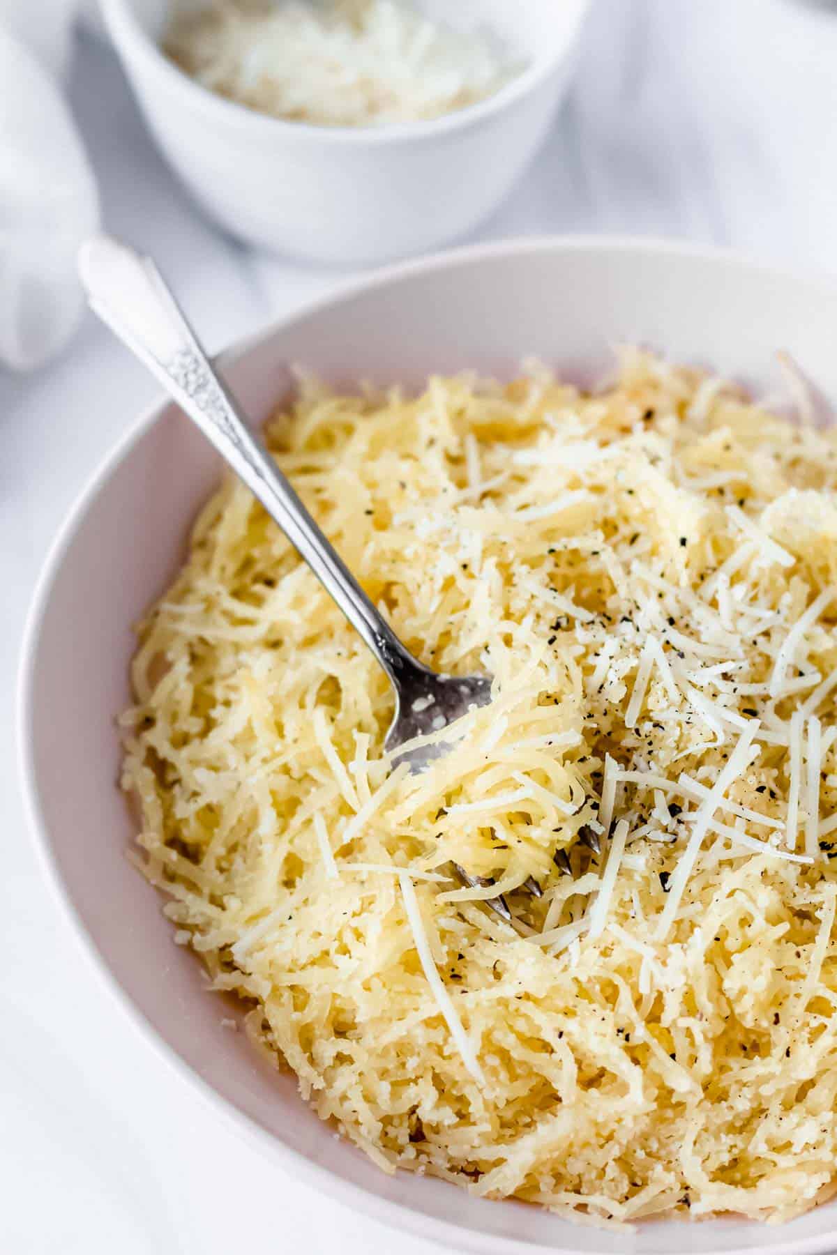 Close up of a pink bowl with spaghetti squash cacio e pepe and a fork in it with a small white bowl of cheese in the background