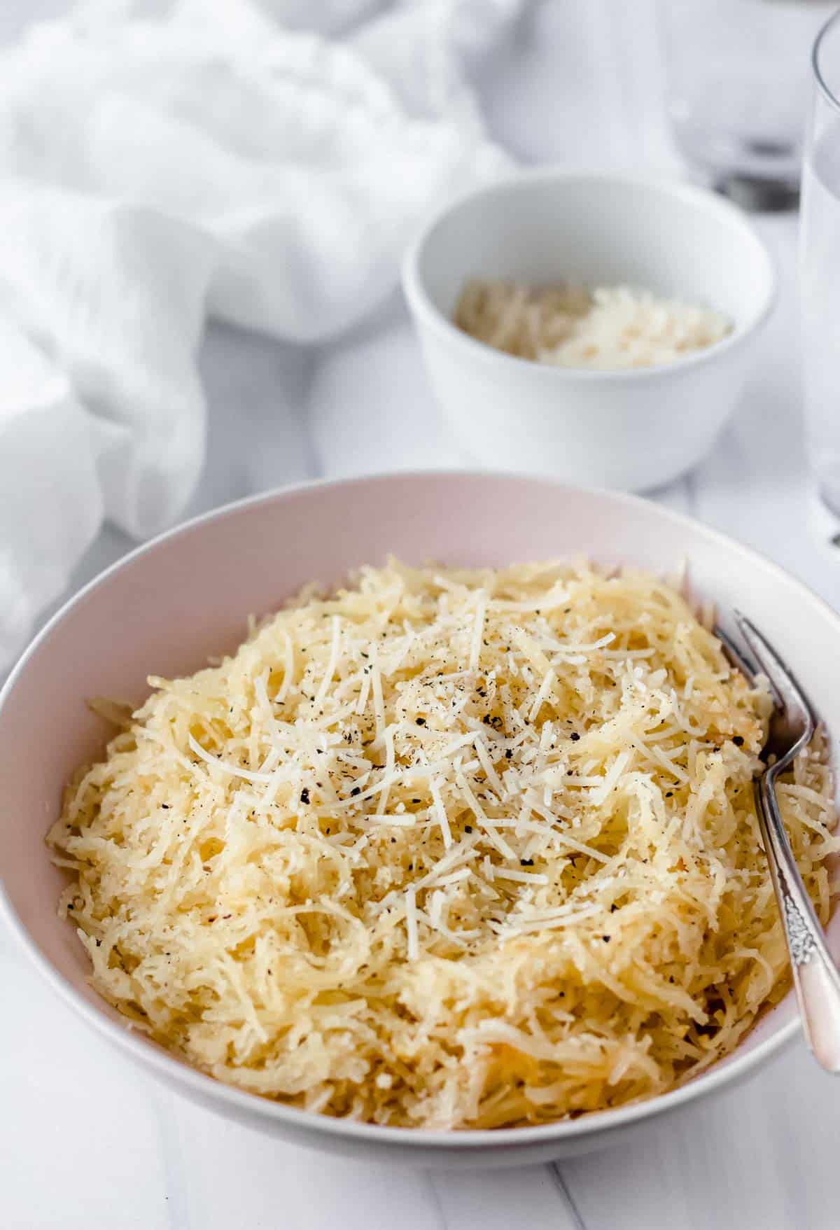 A pink bowl of spaghetti squash cacio e pepe with a fork in it, a small white bowl of cheese, a glass of water and a white towel in the background