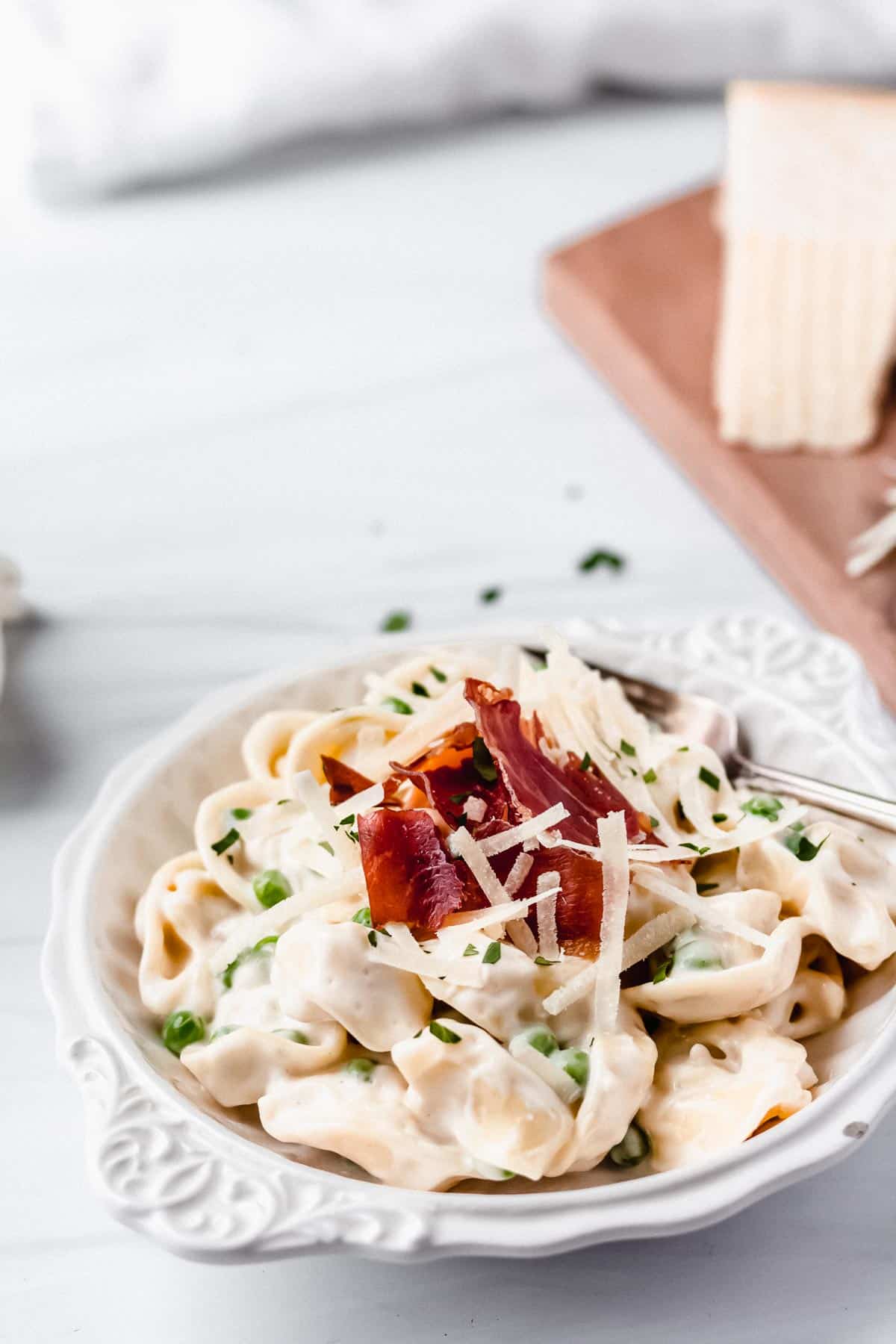 Close up of a white bowl of tortellini alfredo with a cutting board with cheese on it and a white towel in the background