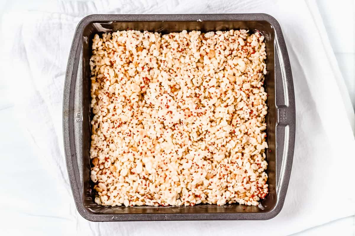 Crispy cereal treats with cranberry seeds pressed into a square baking pan over a white background