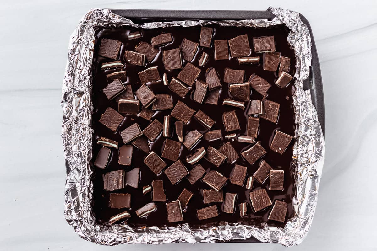 Brownies in a square pan topped with ganache and chopped Andes candies over a white backdrop