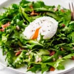 Lyonnaise salad with a poached egg cut open over the top on a white plate over a white background