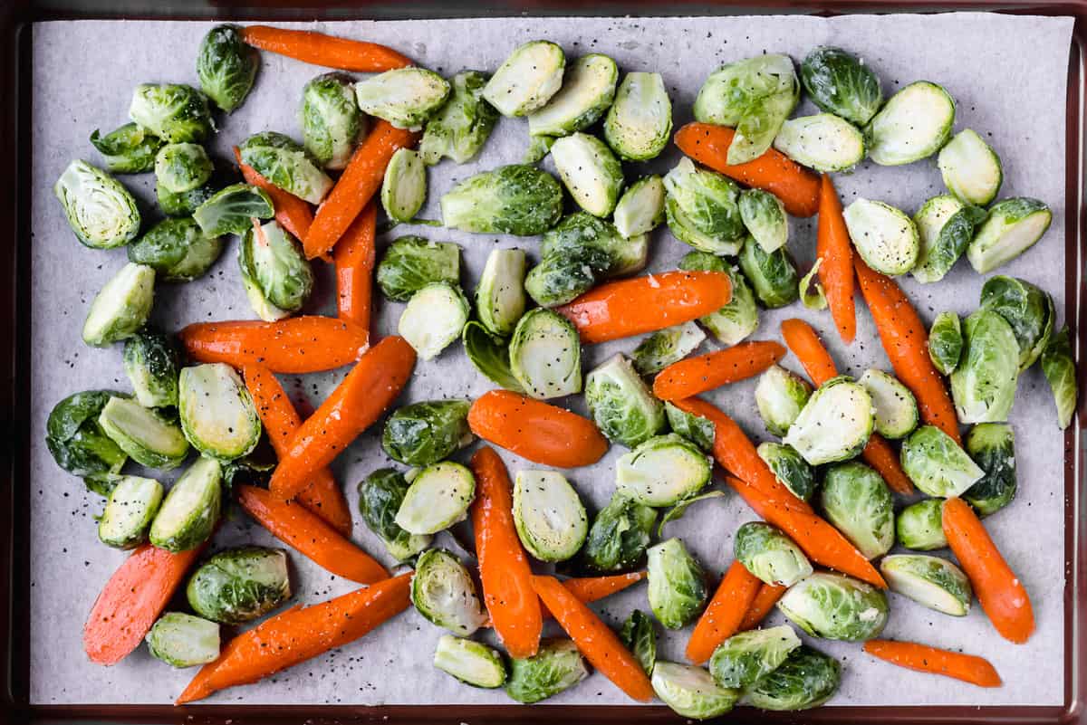Raw brussels sprouts and carrots tossed with maple syrup and butter on a baking sheet lined with parchment paper