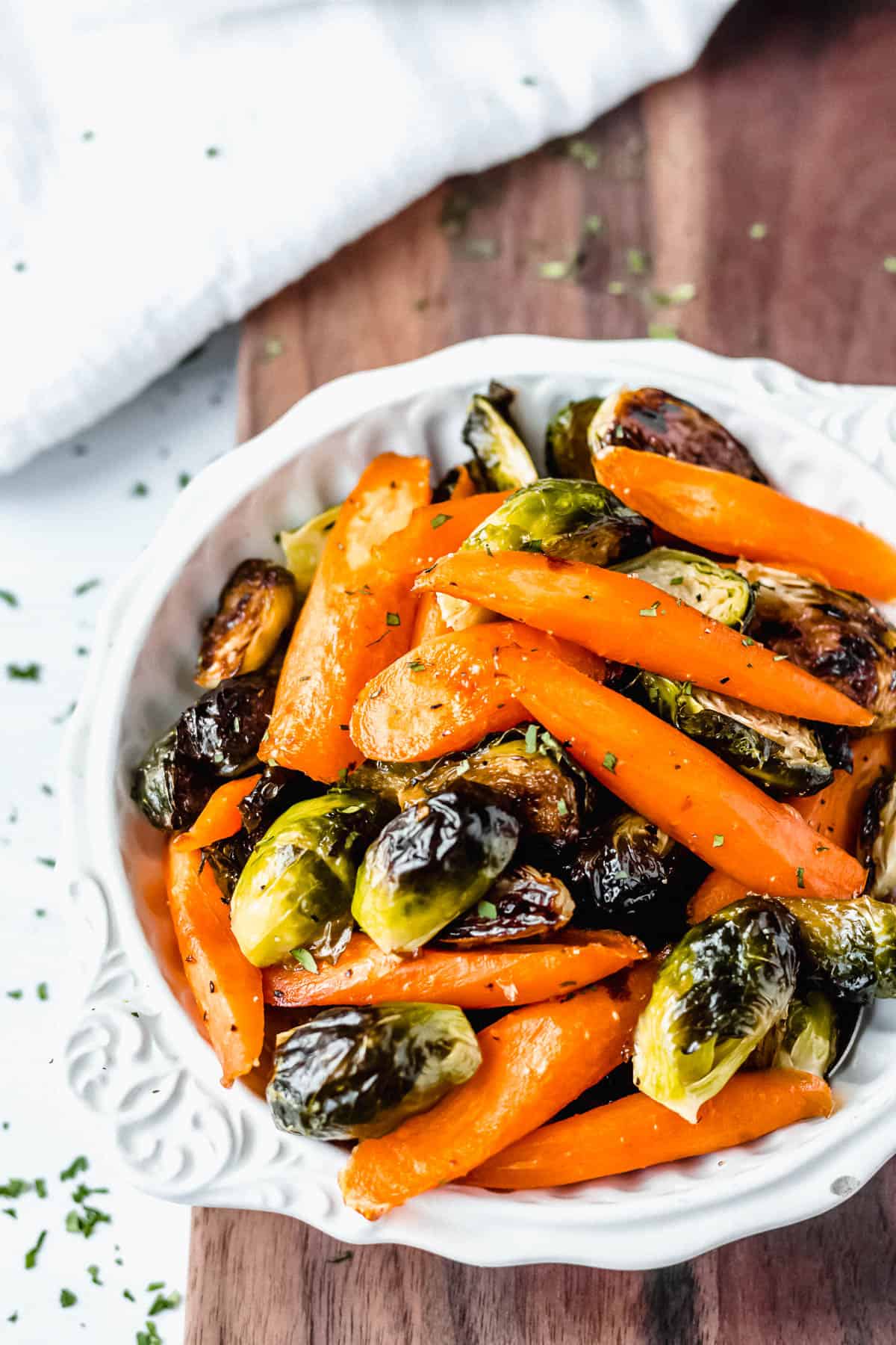 Maple roasted brussels sprouts and carrots in a white bowl over a wood board with a white towel in the background