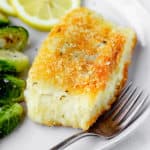 Close up of potato crusted cod on a plate with a fork, brussels sprouts and slices of lemon partially slowing
