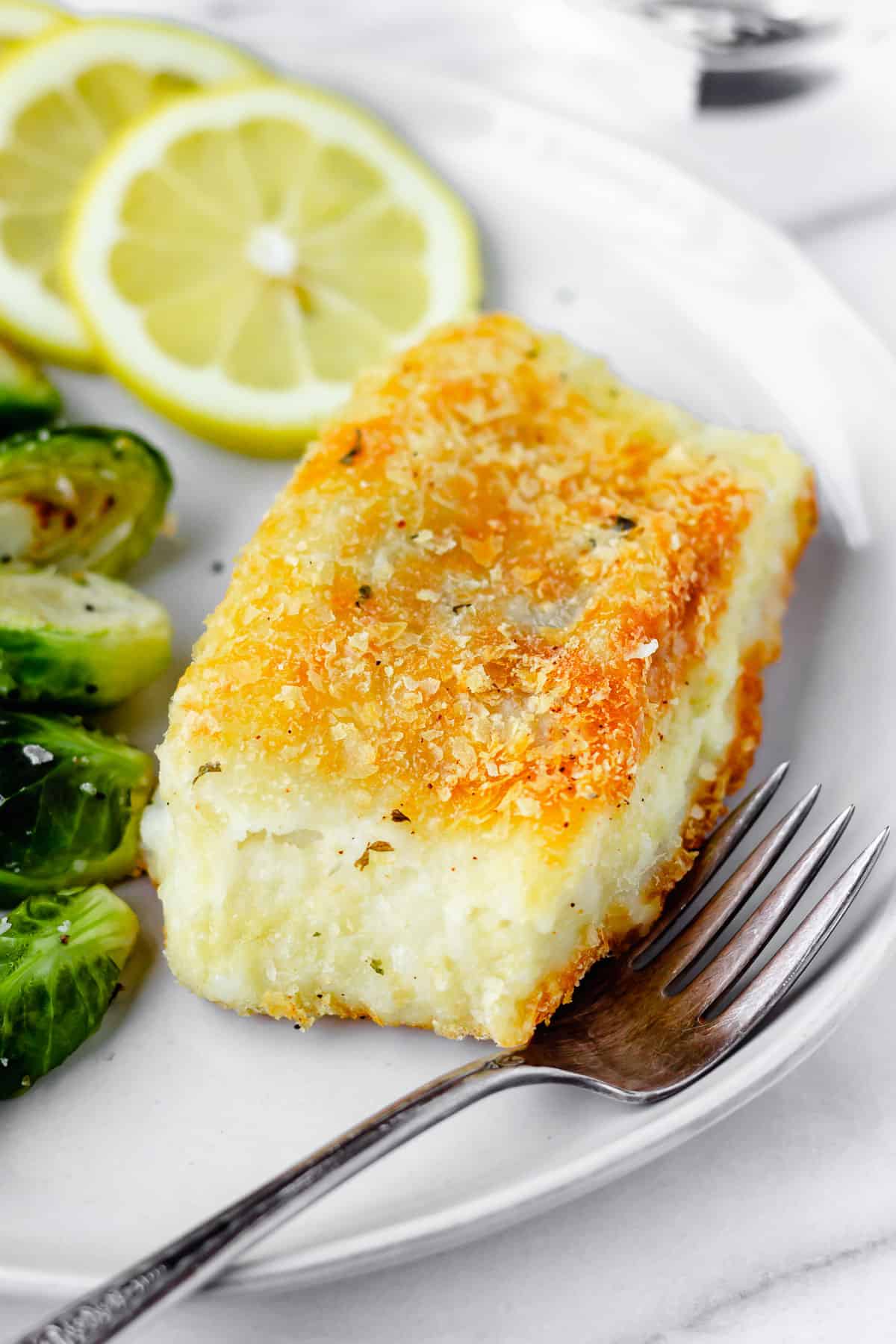 Potato crusted cod on a plate with a fork, brussels sprouts and slices of lemon partially slowing