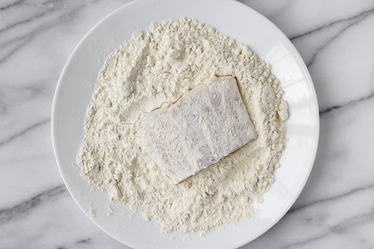 Cod fillet dredges in flour on a white plate over a marble backdrop