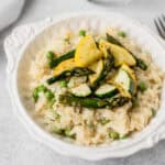 A white bowl filled with risotto and topped with spring vegetables including asparagus, squash and zucchini.
