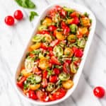 Tomato basil salad in an oval white dish with tomatoes and basil around it on a marble background
