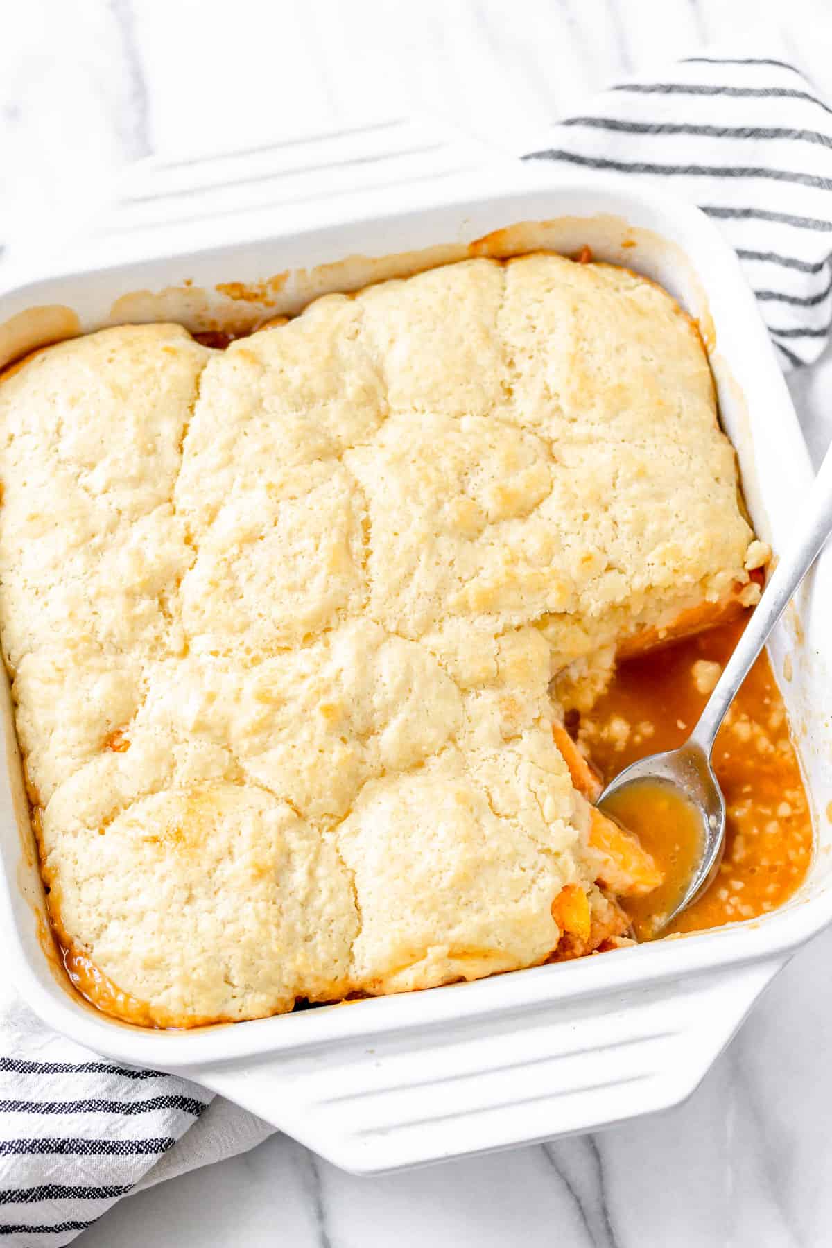 A baking dish of apricot cobbler, with a serving taken out and a spoon inside with juices and slices of fruit