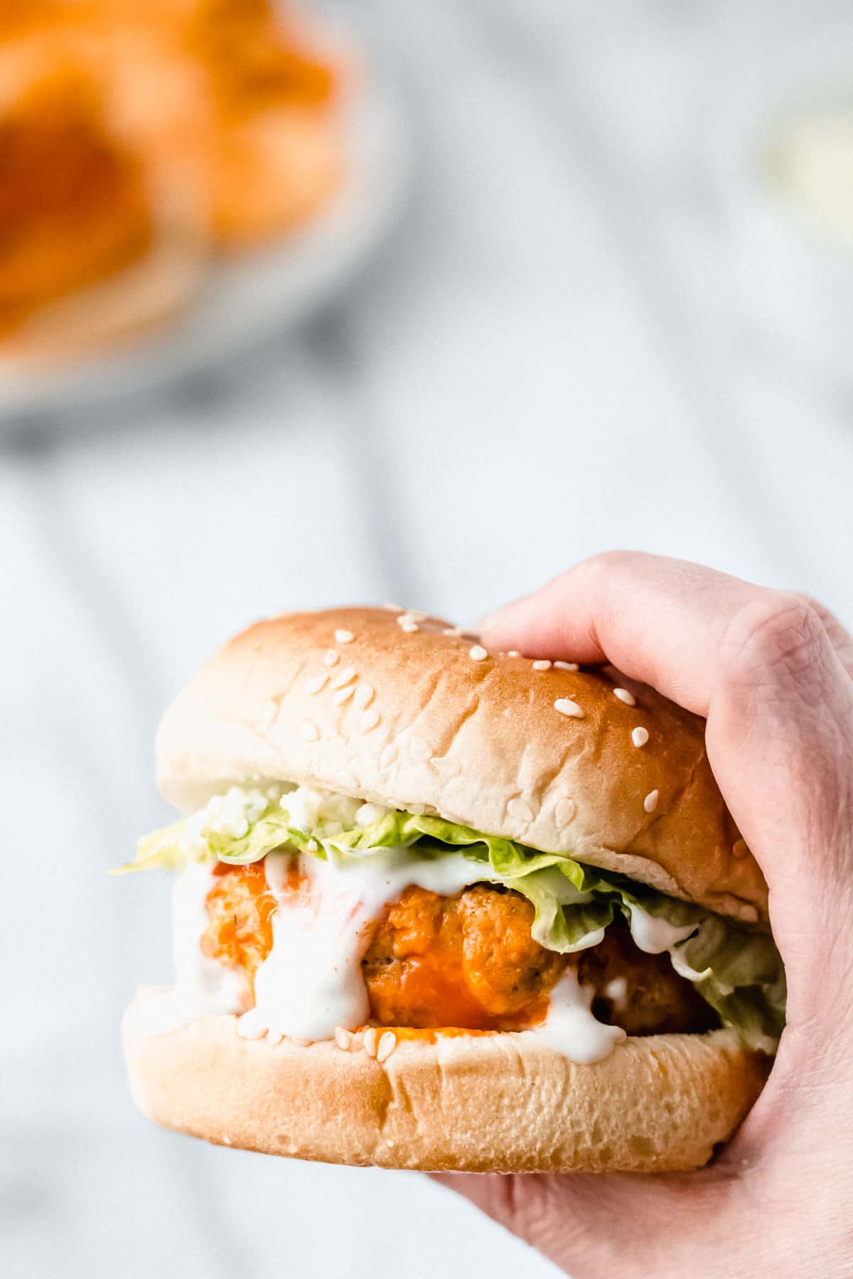 A hand holding a buffalo chicken burger with a second plate blurred in the background