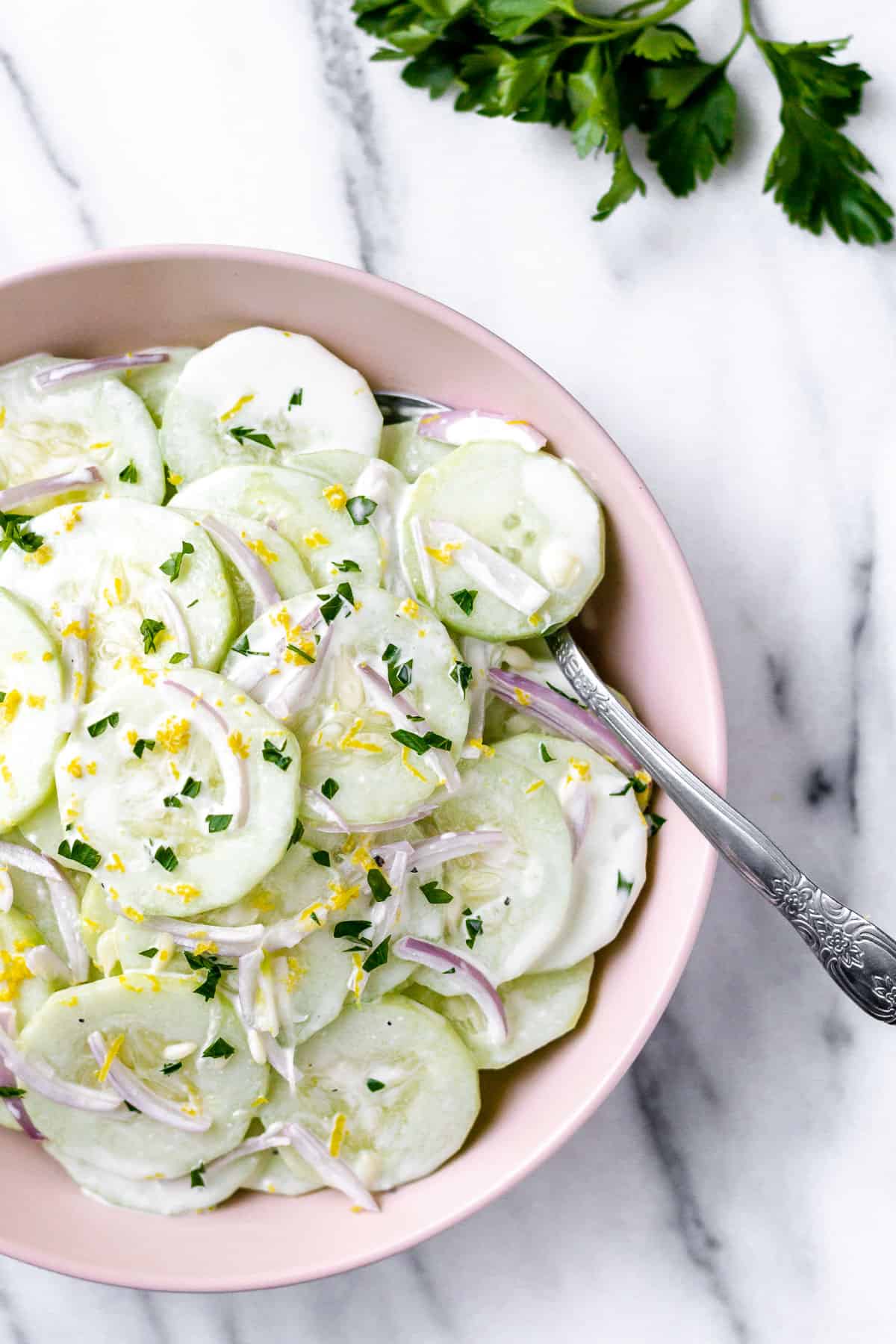 Overhead of a bowl of keto creamy cucumber salad with a spoon in it and fresh parsley in the background