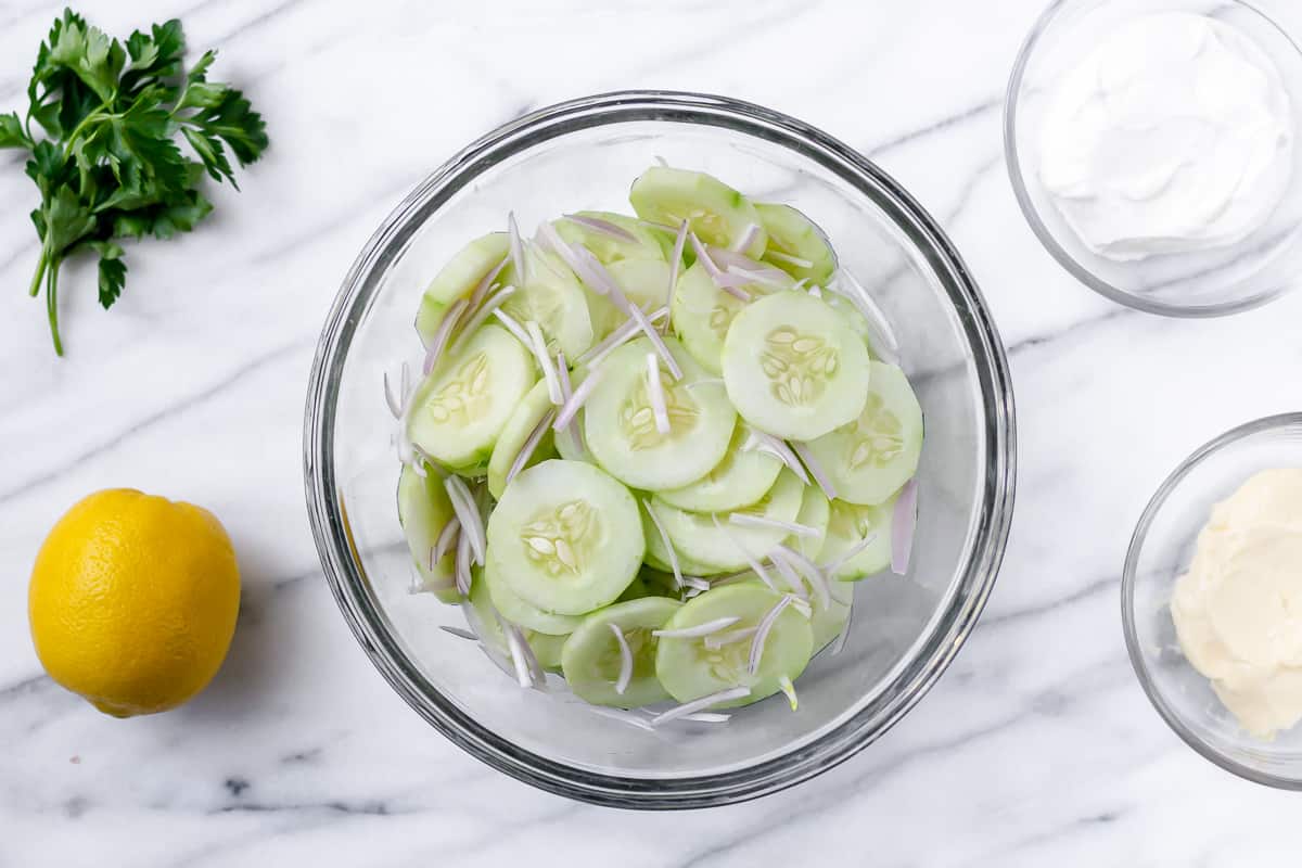 Cucumbers and shallots in a glass bowl with other ingredients around it on a marble backdrop