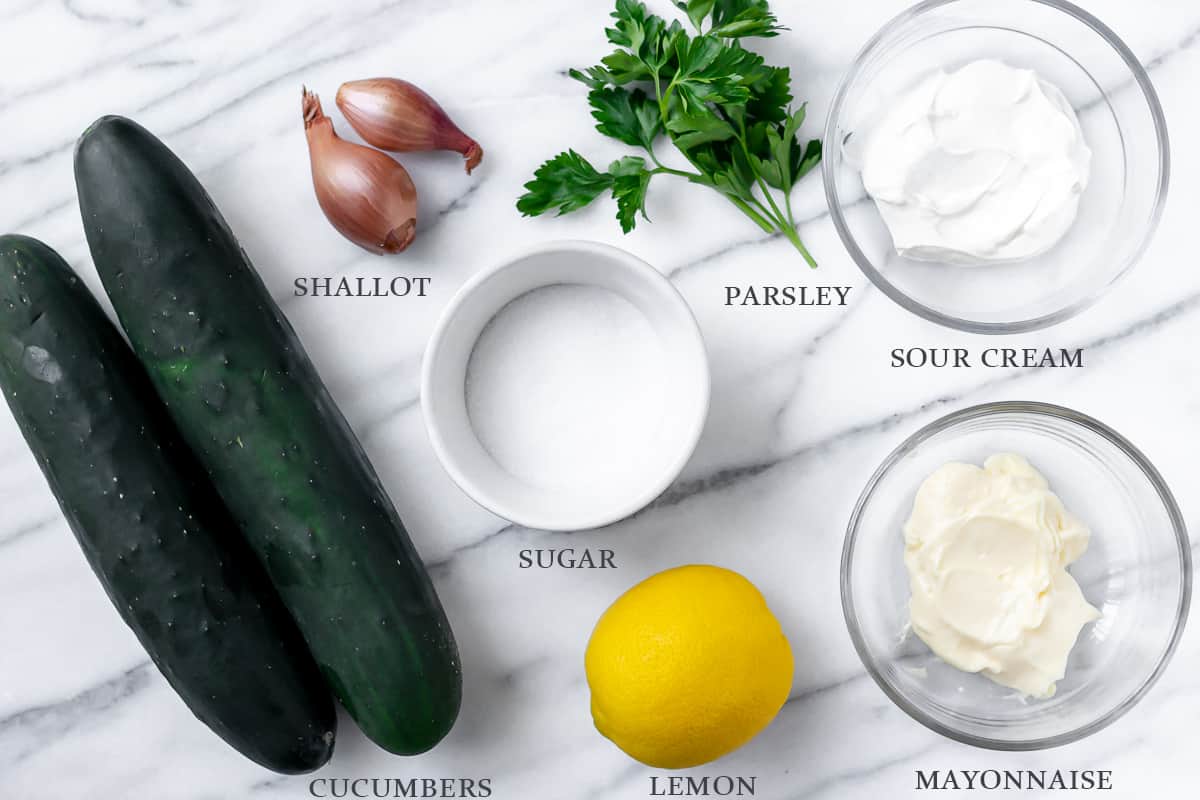 Ingredients needed to make creamy cucumber salad on a marble backdrop with labels