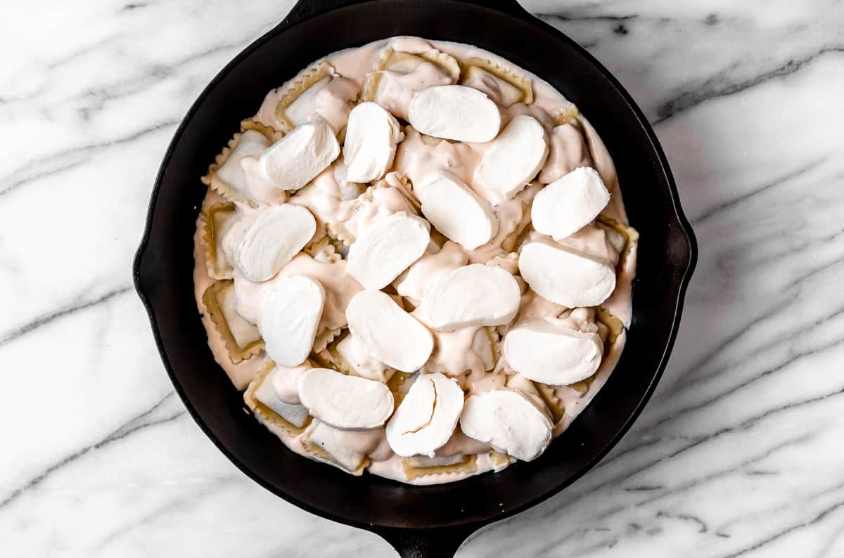 Ravioli topped with alfredo sauce and mozzarella cheese in a cast iron skillet on a marble backdrop